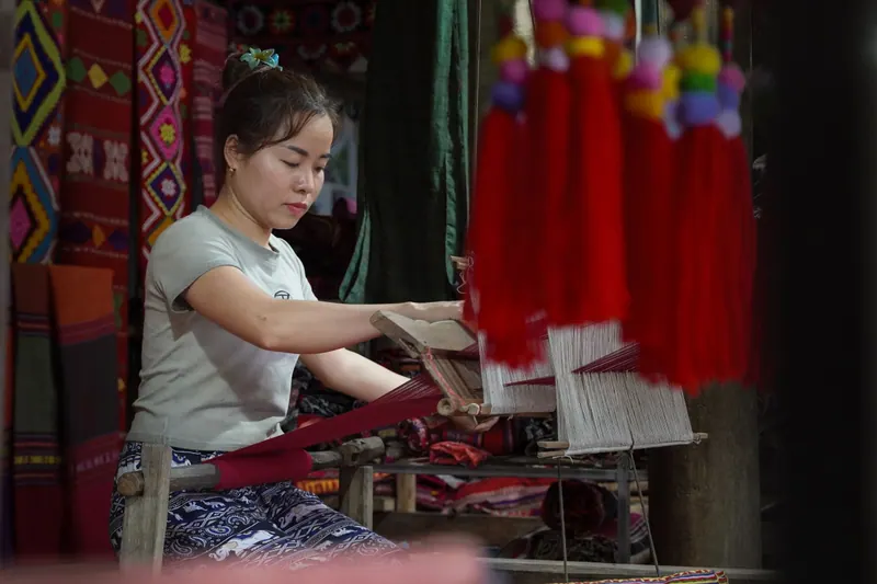 A young weaver at a wooden loom surrounded by red brocade, Thanh Hoa