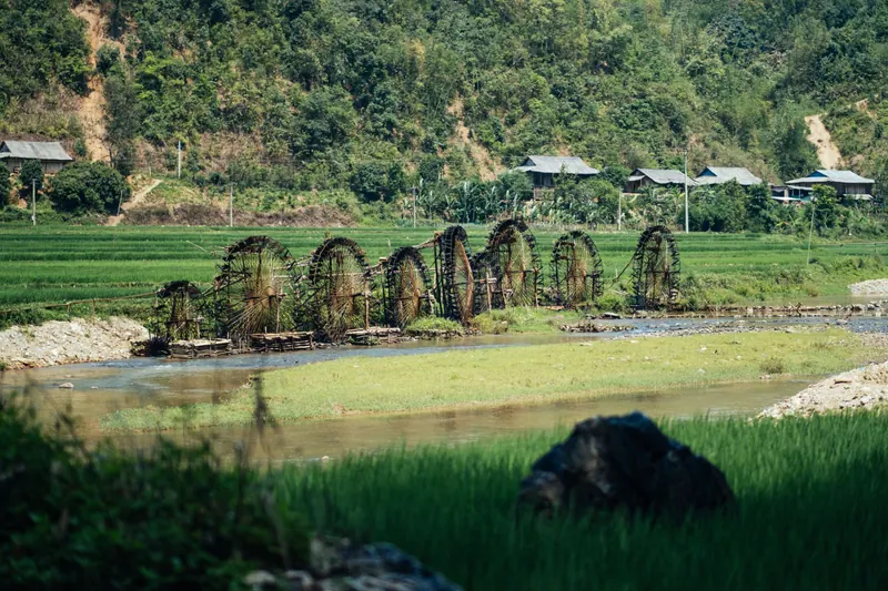 Bamboo water wheels on a stream below rice fields and a stilt-house village